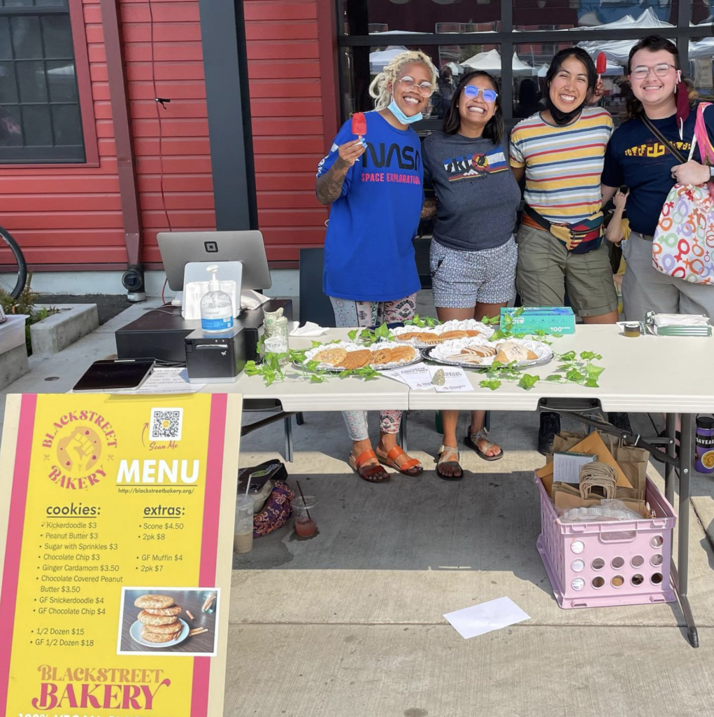 Solidarity Squad volunteers Black Street Bakery pop-up four people smiling at a folding table selling vegan baked goods social justice