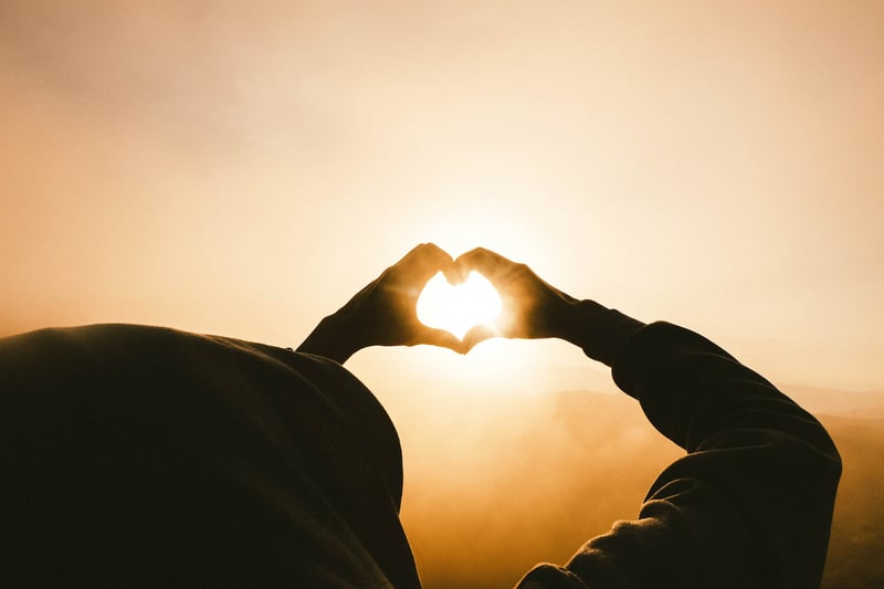 Person making a heart with their hands in front of the sun.
